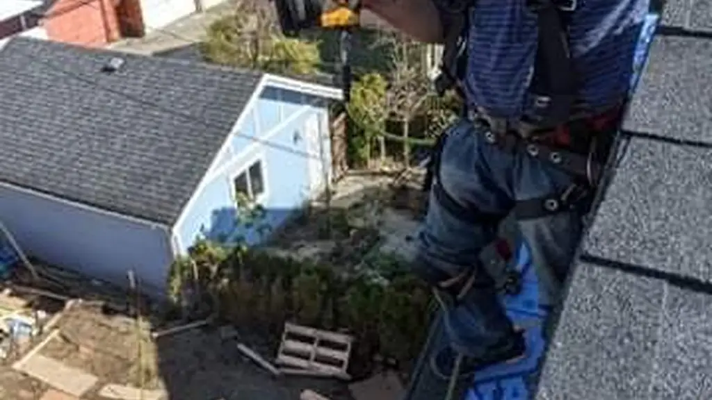 A worker leans over a roof edge smiling directly at the camera while holding a pneumatic nail gun, wearing a full harness and a striped shirt. Fraser Roofing