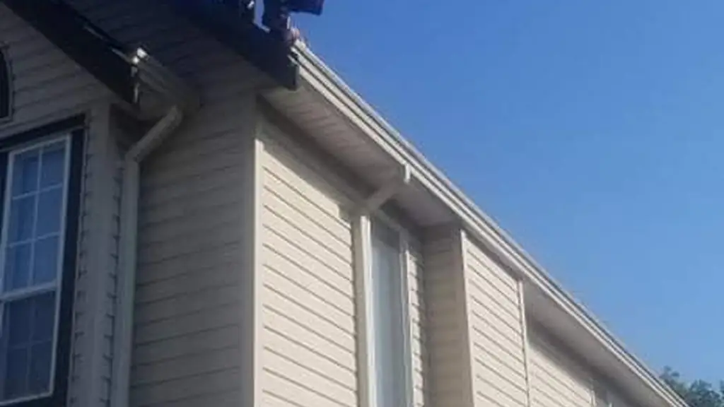 A worker in camouflage clothing stands with both arms outstretched at the ridge of a two-storey house's dark asphalt shingle roof. Fraser Roofing