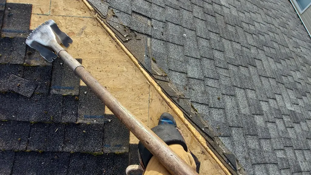 A point-of-view shot looking down at a person's own leg and boot resting at a roof eave, with a tear-off bar laid across the leg. Fraser Roofing